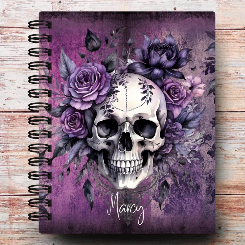 May include: Spiral-bound notebook with a purple floral skull design. The skull is white with intricate details, surrounded by purple roses and dark leaves. The name "Marcy" is written in cursive at the bottom.
