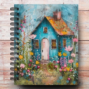 May include: Spiral-bound notebook featuring a whimsical illustration of a blue cottage with an orange roof, surrounded by colorful flowers. The name "Cassandra" is written in cursive at the bottom. The notebook has a wooden background.