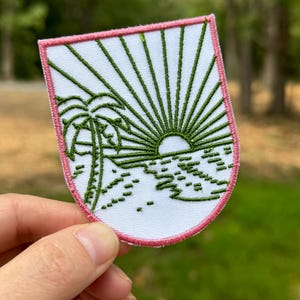 May include: A white embroidered patch with a green palm tree, sun, and ocean waves. The patch has a pink border and is shaped like a shield.