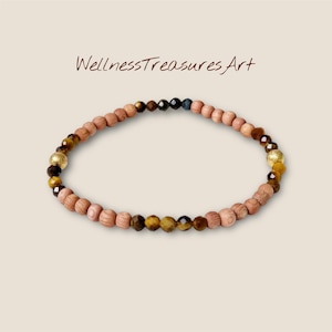 Könnte beinhalten: Ein Perlenarmband mit hellbraunen Holzperlen, Tigerauge, goldfarbenen und dunkelbraunen Perlen. Der Text "WellnessTreasuresArt" befindet sich über dem Armband.