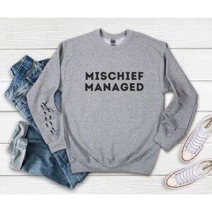 Puede incluir: Una sudadera gris jaspeada con las palabras "MISCHIEF MANAGED" impresas en negro. La sudadera tiene cuello redondo y mangas largas. Huellas negras impresas en la manga izquierda. Vaqueros azules y zapatillas blancas en el fondo.