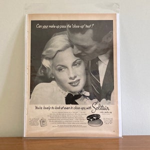 May include: A black and white vintage advertisement for Solitair cake makeup. The ad features a couple in a close-up shot with the text "Can your make-up pass the 'close-up' test?" and "You're lovely-to-look-at even in close ups, with Solitair cake make-up."