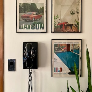 May include: A wall-mounted display featuring framed vintage advertisements and a black rotary dial telephone. The ads showcase a red Datsun car and colorful outdoor carpet. A black light switch is also visible.