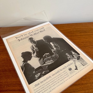 May include: Vintage advertisement for Johnnie Walker Red Scotch whisky. The black and white image features people enjoying the drink. Text includes "You'll be glad you said Johnnie Walker Red" and details about the whisky.