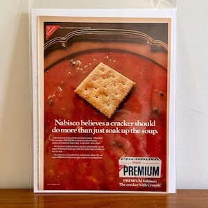 May include: A vintage advertisement for Nabisco Premium Saltines crackers. The ad features a single cracker floating in a bowl of tomato soup. The text reads "Nabisco believes a cracker should do more than just soak up the soup." and "PREMIUM Saltines. The cracker with Crunch."