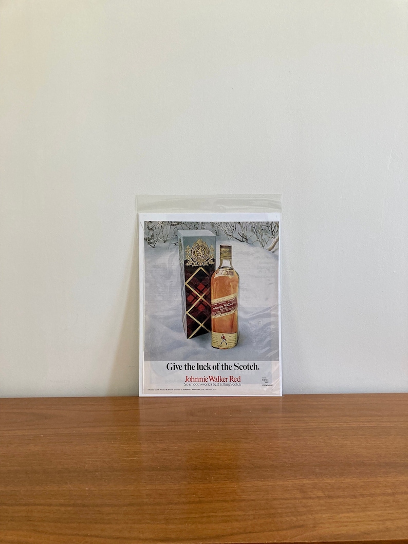 May include: A vintage print advertisement for Johnnie Walker Red Scotch whisky. The ad features a bottle of Johnnie Walker Red Scotch whisky in a snowy setting with the text "Give the luck of the Scotch. Johnnie Walker Red. So smooth-world's best selling Scotch."