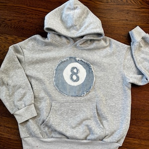 Puede incluir: Una sudadera gris con bolsillo delantero y capucha. La sudadera presenta un parche circular con el número 8 en blanco, sobre un fondo de mezclilla azul. La sudadera tiene mangas largas y un corte holgado.