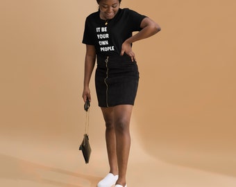 Puede incluir: Una mujer con una camiseta negra con el texto "IT BE YOUR OWN PEOPLE" en letras blancas. También lleva una falda negra y zapatos blancos de vestir.