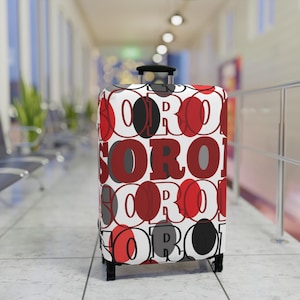 May include: A rolling suitcase with a repeating pattern of the word "SORO" in red, black, and gray on a white background. The suitcase has a black handle and wheels, and is in an airport setting.