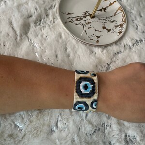 May include: White beaded bracelet with a pattern of blue evil eyes. The bracelet is made of small, round beads and has a woven design.
