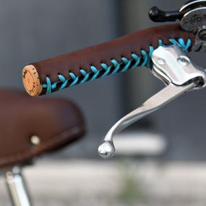 May include: Close-up of a bicycle handlebar with a brown leather grip and turquoise stitching. The end of the grip has a cork stopper. A silver brake lever is also visible, along with a brown leather bicycle seat.