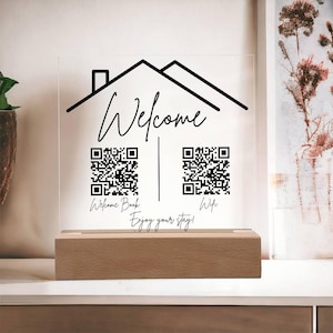 May include: A white acrylic sign with a black house outline and the word "Welcome" in cursive. The sign has two black and white QR codes, one labeled "Welcome Book" and the other labeled "Wifi". The sign is sitting on a wooden base. The text "Enjoy your stay!" is written below the QR codes.