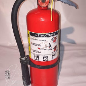 May include: A red fire extinguisher with a black hose and handle. The extinguisher has a pressure gauge and instructions label. The text "INSTRUCTIONS" is visible on the label. The extinguisher is labeled "AMEREX 5 LBS 2 CHAMBER".