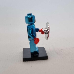 Puede incluir: Una figura de Lego azul met&aacute;lico que sostiene un escudo plateado y rojo. La figura tiene guantes de boxeo rojos y est&aacute; de pie sobre una base negra. Es un juguete o art&iacute;culo de colecci&oacute;n.