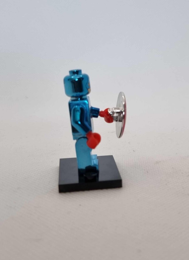 Puede incluir: Una figura de Lego azul met&aacute;lico que sostiene un escudo plateado y rojo. La figura tiene guantes de boxeo rojos y est&aacute; de pie sobre una base negra. Es un juguete o art&iacute;culo de colecci&oacute;n.