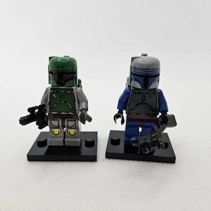 May include: Two Lego Star Wars minifigures. One figure is in green and grey armour with a black weapon. The other figure is in blue and grey armour with a black weapon. Both figures stand on black bases.