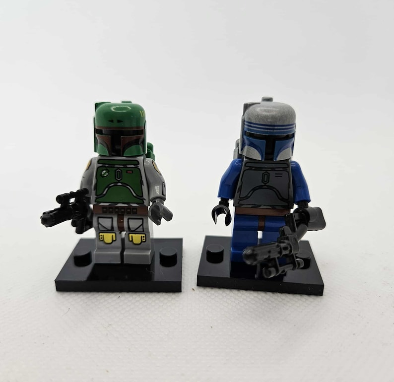 K&ouml;nnte beinhalten: Zwei Lego Star Wars Minifiguren. Eine Figur tr&auml;gt eine gr&uuml;ne und graue R&uuml;stung mit einer schwarzen Waffe. Die andere Figur tr&auml;gt eine blaue und graue R&uuml;stung mit einer schwarzen Waffe. Beide Figuren stehen auf schwarzen Sockeln.