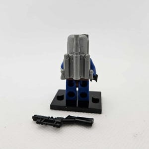 May include: A Lego minifigure of a character from Star Wars, viewed from the back. The figure wears a blue suit, gray backpack, and a black base. A black blaster rifle lies in front of the figure.