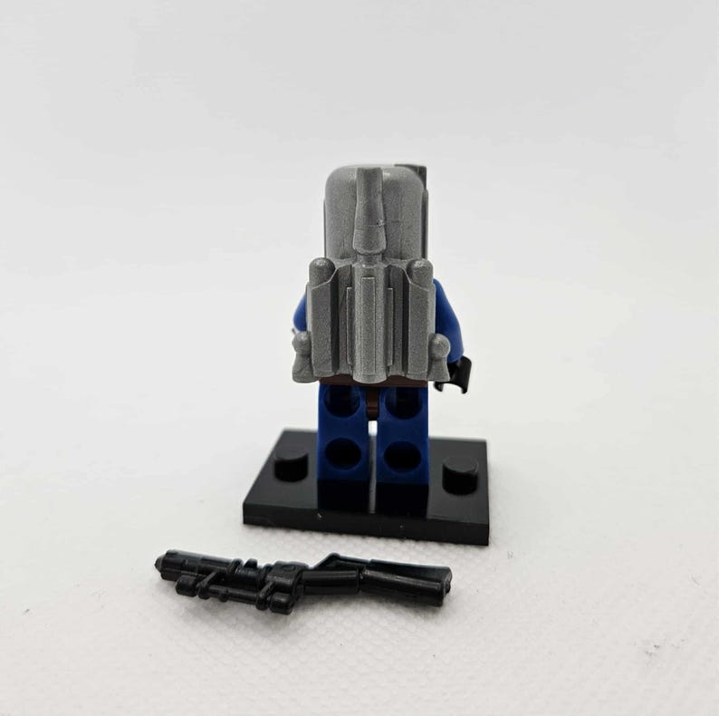 May include: A Lego minifigure of a character from Star Wars, viewed from the back. The figure wears a blue suit, gray backpack, and a black base. A black blaster rifle lies in front of the figure.
