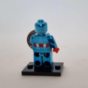 May include: A metallic blue Lego minifigure of a superhero, with red and white accents on the torso and red gloves. The figure is standing on a black base and holding a silver disc. The overall theme is collectibles.