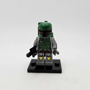 K&ouml;nnte beinhalten: Eine Lego Star Wars Boba Fett Minifigur. Die Figur hat einen gr&uuml;nen Helm und R&uuml;stung, graue Arme und Beine und einen schwarzen Blaster. Sie steht auf einer schwarzen Basis.