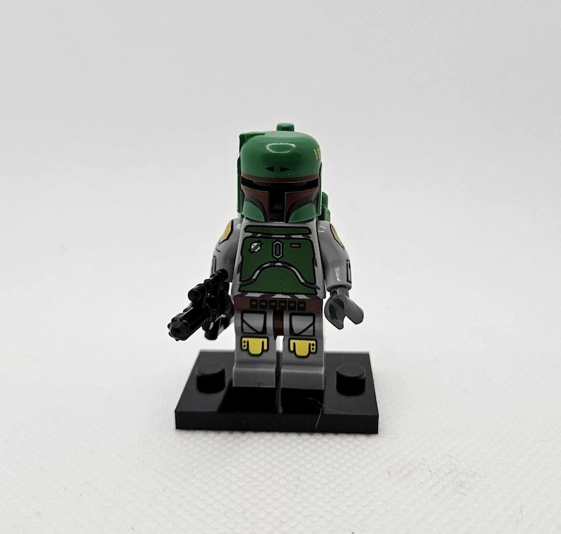 K&ouml;nnte beinhalten: Eine Lego Star Wars Boba Fett Minifigur. Die Figur hat einen gr&uuml;nen Helm und R&uuml;stung, graue Arme und Beine und einen schwarzen Blaster. Sie steht auf einer schwarzen Basis.
