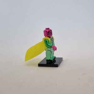 Puede incluir: Una figura de acci&oacute;n verde met&aacute;lico con cabeza y manos rosas, y una capa amarilla. La figura se encuentra sobre una base negra. El dise&ntilde;o de la figura recuerda a un superh&eacute;roe.