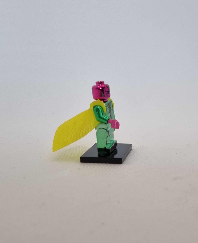 Puede incluir: Una figura de acci&oacute;n verde met&aacute;lico con cabeza y manos rosas, y una capa amarilla. La figura se encuentra sobre una base negra. El dise&ntilde;o de la figura recuerda a un superh&eacute;roe.
