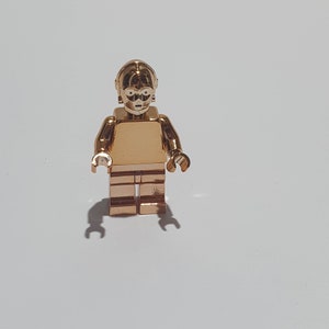 Puede incluir: Una minifigura Lego de C-3PO, un personaje de la franquicia Star Wars, de color dorado. La figura está de pie con los brazos a los lados.