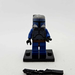 K&ouml;nnte beinhalten: Eine Lego-Minifigur eines Charakters mit blauem und grauem Helm und R&uuml;stung, mit blauen Beinen und Armen. Die Figur steht auf einer schwarzen Basis, und ein schwarzes Blastergewehr liegt davor. Die Figur ist ein Star Wars-Charakter.