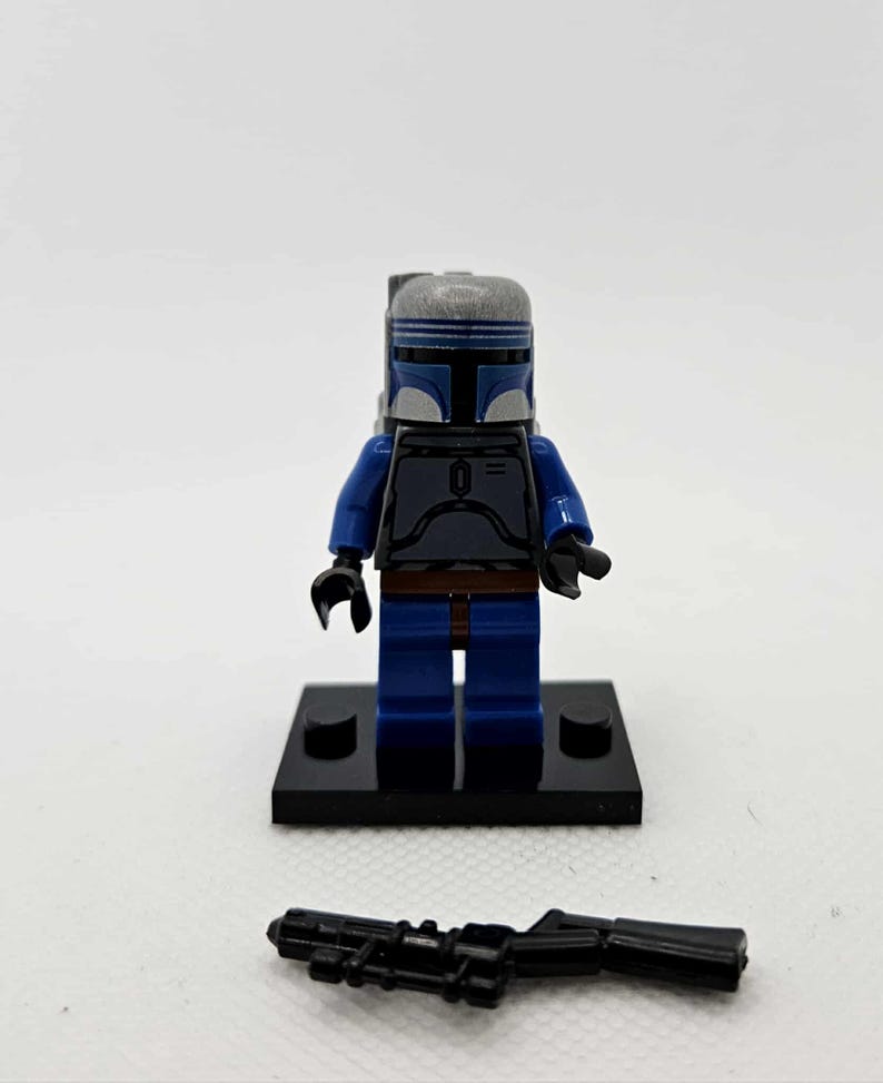 K&ouml;nnte beinhalten: Eine Lego-Minifigur eines Charakters mit blauem und grauem Helm und R&uuml;stung, mit blauen Beinen und Armen. Die Figur steht auf einer schwarzen Basis, und ein schwarzes Blastergewehr liegt davor. Die Figur ist ein Star Wars-Charakter.