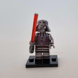 May include: A metallic silver Darth Vader Lego figure with a red lightsaber. The figure is standing on a black base. The helmet and armor have a reflective finish, and the lightsaber is held in the right hand.