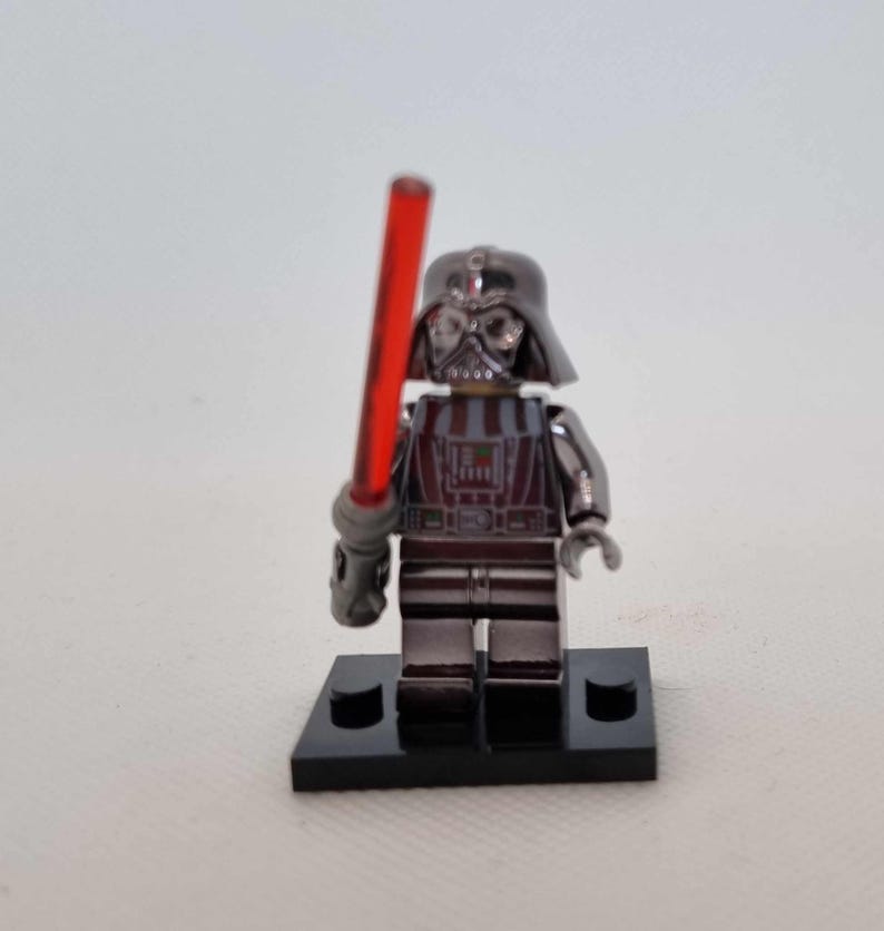 May include: A metallic silver Darth Vader Lego figure with a red lightsaber. The figure is standing on a black base. The helmet and armor have a reflective finish, and the lightsaber is held in the right hand.
