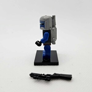 May include: A Lego minifigure of a character in a gray helmet with blue accents, blue arms and legs, and a gray backpack. The figure stands on a black base, with a black blaster rifle in front.