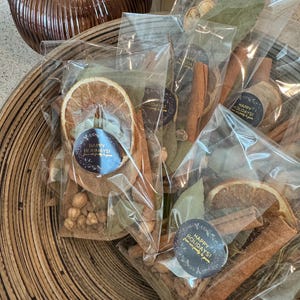 Może przedstawiać: Kilka przezroczystych torebek celofanowych wypełnionych suszonymi plasterkami pomarańczy, laskami cynamonu, liśćmi laurowymi i przyprawami. Każda torebka ma naklejkę z napisem "Happy Holidays!". Torebki ułożone są w drewnianej misce.