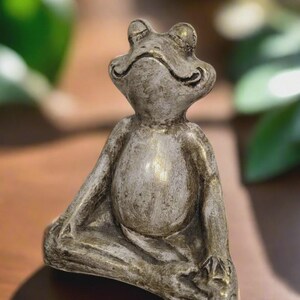 May include: A small, silver-colored frog statue in a seated yoga pose. The frog has a serene expression and is detailed with textured lines. The statue is likely made of resin or a similar material, and is suitable for indoor or outdoor decor.