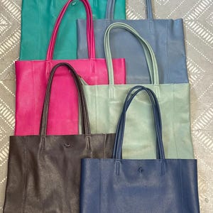May include: A collection of leather tote bags in various colors, including teal, pink, gray, and navy. Each bag has a matching set of handles and a subtle logo. The bags are arranged in a neat, stacked display.