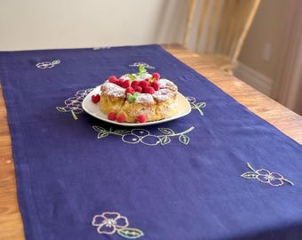 Può includere: Un runner da tavola rettangolare blu navy con motivi floreali ricamati in filo bianco e verde. Il tessuto presenta una trama visibile e il ricamo è distribuito sulla superficie in un motivo floreale che si ripete.