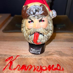 Könnte beinhalten: Eine Krampus-Figur mit roter Weihnachtsmütze, langem weißem Bart und roter Zunge. Das Wort "Krampus" ist in roter Schreibschrift auf einer Holzoberfläche geschrieben. Die Figur steht auf einem schwarzen Sockel.