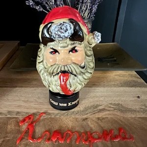 Könnte beinhalten: Ein Keramik-Weihnachtsmannkopf mit roter Mütze und wütendem Gesichtsausdruck. Die Santa-Figur hat einen langen weißen Bart und eine rote Zunge, die herausragt. Auf der Basis der Figur steht der Text "Gruss vom Krampus".