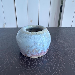 May include: A small, round ceramic vase with a wide opening. The vase has a light blue and beige glaze with hints of pink near the base. The handmade pottery sits on a dark brown surface with a floral pattern, against a white wooden background.