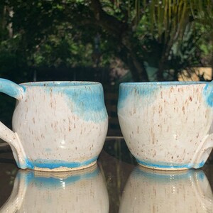 Peut inclure: Deux tasses en céramique blanche avec un glaçage bleu et des mouchetures brunes. Les tasses ont un corps arrondi et une anse.