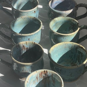 May include: A collection of ten teal blue ceramic mugs with brown speckles and a glazed finish. The mugs have rounded handles and are arranged in a staggered pattern.