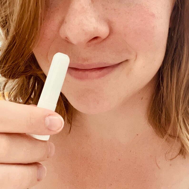 May include: A white, cylindrical lip balm tube is held near a person's lips. The tube is smooth and appears to be made of plastic. The person's skin is fair with a slight blush.