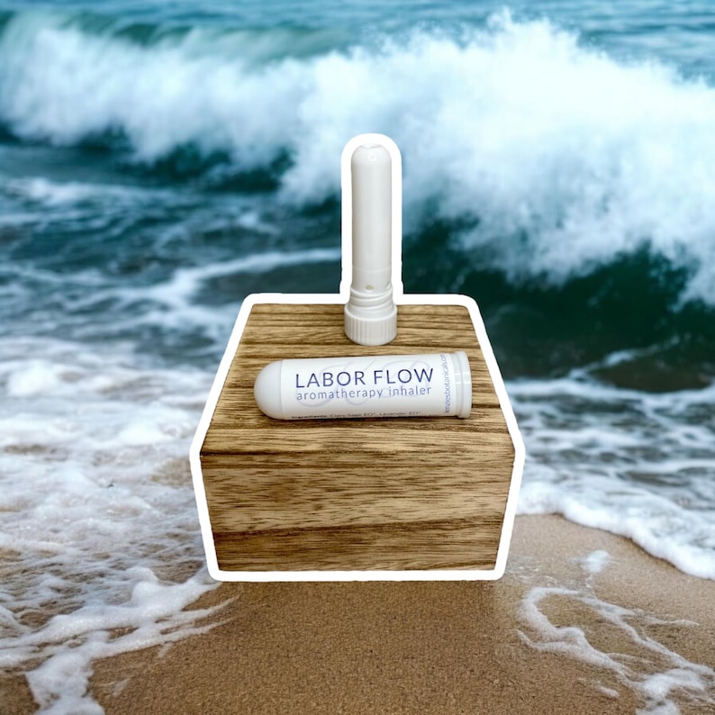 May include: Two white aromatherapy inhalers, one assembled and one disassembled, are displayed on a wooden block. The assembled inhaler stands upright. The other inhaler is horizontal, with the text "LABOR FLOW aromatherapy inhaler". The background features a beach and ocean.