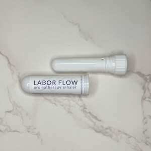 May include: Two white aromatherapy inhalers, one assembled and one disassembled, are displayed on a marble surface. The assembled inhaler has the text "LABOR FLOW aromatherapy inhaler" printed on it. The disassembled inhaler has a screw-on cap.