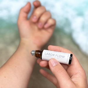 May include: A small brown glass bottle with a rollerball applicator. The bottle is labeled "LABOR FLOW essential oil roll on" and has a white label with black text. The bottle is being held in a person's hand.