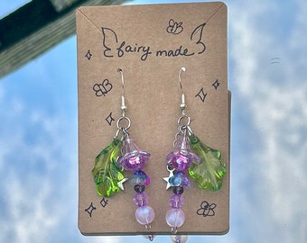 May include: A pair of earrings with a green leaf and purple flower charm, with purple and clear beads hanging below. The earrings are on a brown card with the text "fairy made" written on it.