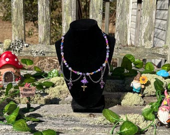 May include: A beaded necklace with purple, blue, and pink beads, displayed on a black mannequin. The necklace has dangling charms and a small cross pendant. The background includes fairy garden decorations and greenery.