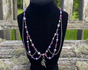 May include: A layered necklace with a silver chain and purple and iridescent beads. The necklace has a silver charm of a crescent moon with a fairy on it.